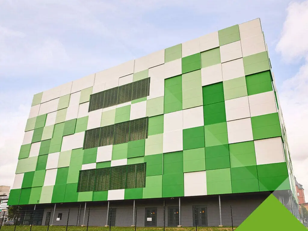 Bild Modernes Gebäude mit einer Fassade aus weißen und grünen geometrischen Paneelen. Mehrere horizontale, rechteckige Fenster sind mittig ausgerichtet. Das Gebäude ist von einem Zaun umgeben und der Himmel ist klar mit ein paar Wolken.
