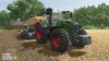 Ein Traktor zieht unter klarem Himmel landwirtschaftliche Geräte durch ein Feld. In einer ländlichen Gegend mit Bäumen und einer Scheune im Hintergrund fängt das Bild das immersive Erlebnis des Landwirtschafts-Simulator 25 Servers perfekt ein.